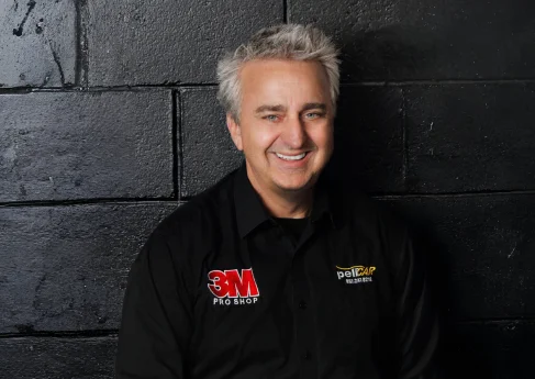 Smiling man in a 3M branded shirt, sitting against a black wall.