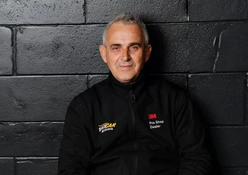 Older man in a black 3M jacket, sitting in front of a black wall.