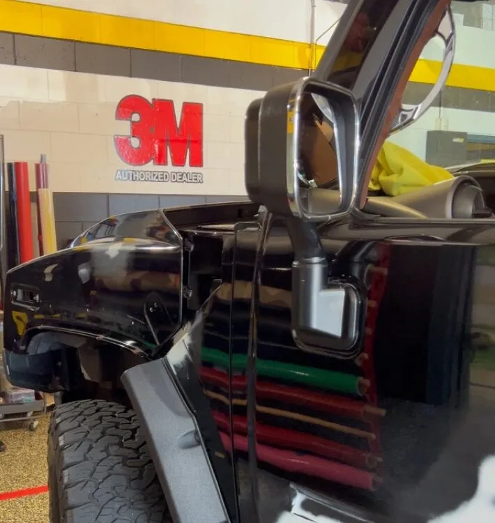 A close-up of a black vehicle with a visible side mirror and a 3M Authorized Dealer sign on the wall behind it.