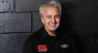 A person wearing a 3M Pro Shop and Pelicar shirt smiling in front of a dark wall.