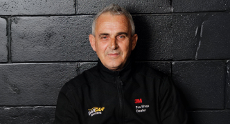 A person with short gray hair, wearing a 3M Pro Shop and Pelicar jacket, sitting against a black wall.