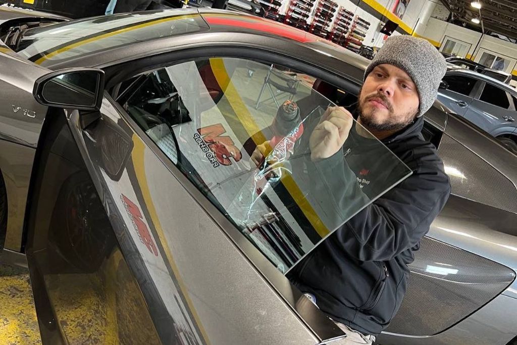 A technician wearing a beanie and black jacket installs window tint film on the driver-side window of a gray Audi R8 V10 inside a professional tinting shop. Rolls of film and a large 3M Pro Shop banner are visible in the background, emphasizing a high-end tinting service.
