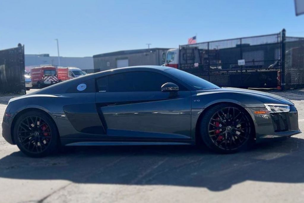 Sleek dark gray Audi R8 with black wheels and heavily tinted windows parked in an industrial lot on a sunny day. The tint is noticeably dark, illustrating a style that may push the limits of Massachusetts' 35% window tint law.