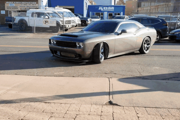 Looping GIF of a sleek gray Dodge Challenger with heavily tinted windows pulling out of a parking lot on a sunny day. The vehicle’s windows reflect the sky, emphasizing the deep tint, while surrounding businesses and inspection stations in Massachusetts are visible in the background.