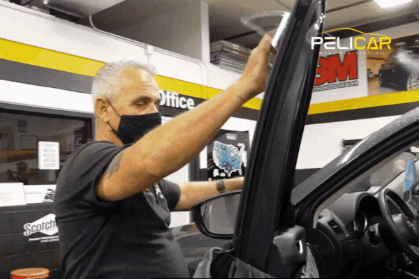 Animated GIF of a masked technician at Pelicar Window Tinting carefully applying film to a car window in a professional garage setting. Branded elements like the Pelicar logo and 3M Pro Shop signage are visible in the background, emphasizing trust and certification.