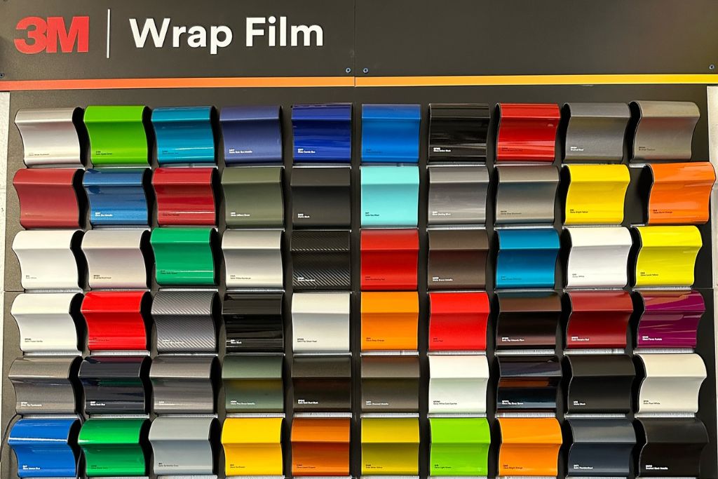 Display wall featuring a wide array of 3M wrap film color samples, including glossy, matte, metallic, satin, and textured finishes in vibrant hues like red, blue, green, yellow, and chrome.