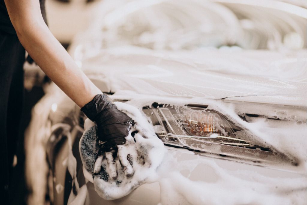 Close-up of a person wearing a black glove hand-washing a soapy car headlight with a microfiber mitt. Thick suds cover the front of the vehicle, indicating gentle cleaning to preserve a vinyl wrap.