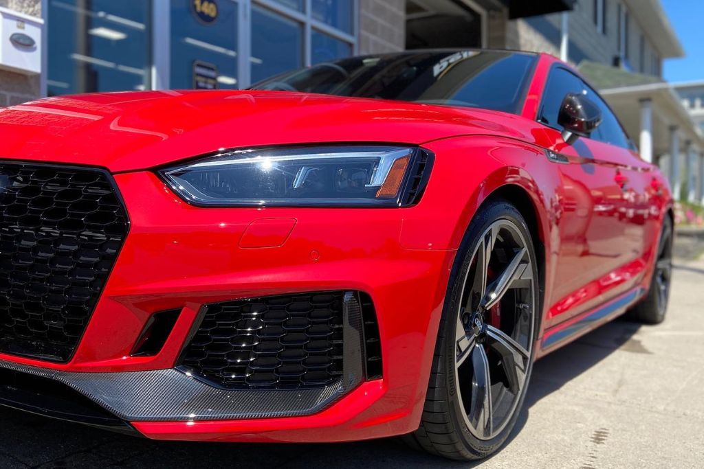 Close-up of a bright red sports sedan with a glossy vinyl wrap, parked outside a commercial building. The wrap highlights the car’s sleek lines while protecting the factory paint beneath.