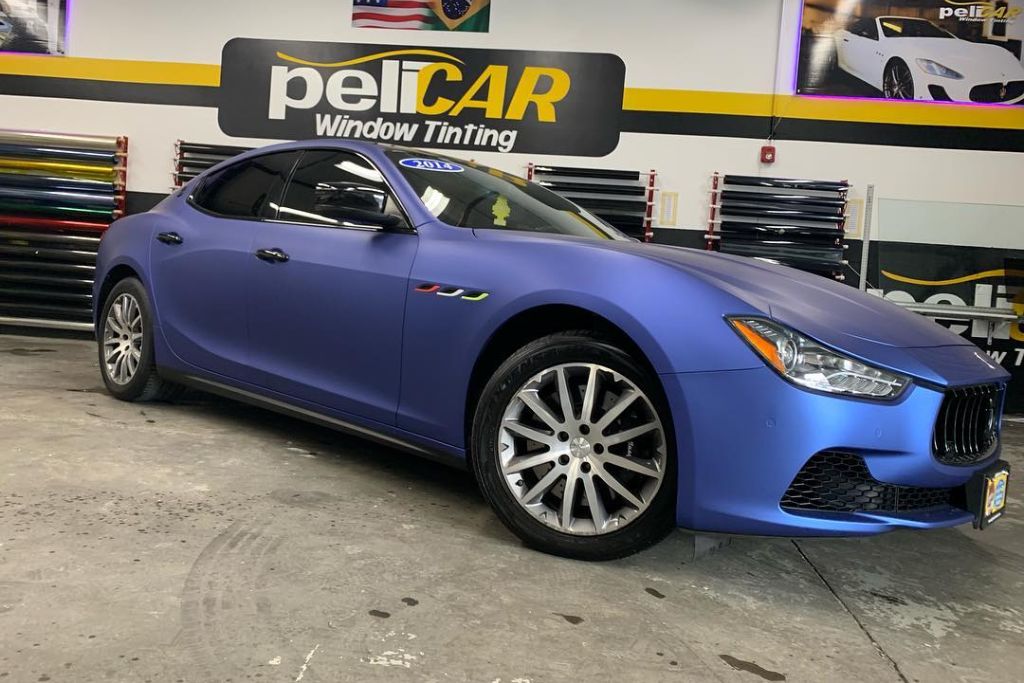 A luxury sedan with a matte blue vinyl wrap is parked inside the Pelicar shop, surrounded by rolls of wrap film and branded signage. The finish appears smooth and flawless, highlighting the wrap’s premium durability.
