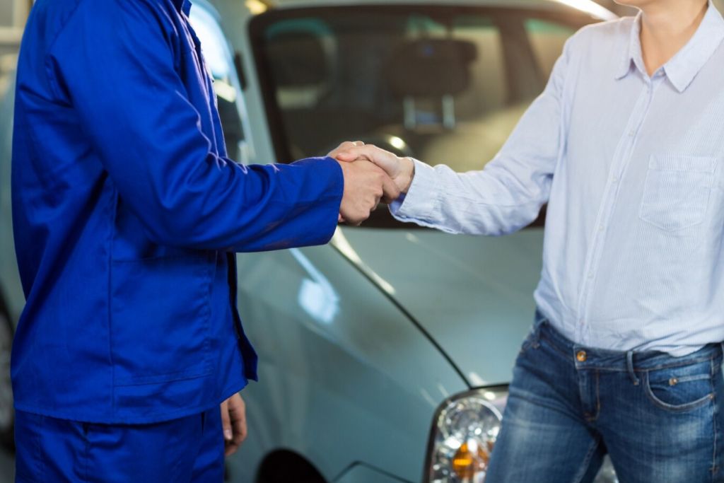 A customer and an auto technician shake hands in front of a silver vehicle, symbolizing a successful service or sale. The technician wears a blue uniform, while the customer is dressed casually in a light shirt and jeans.