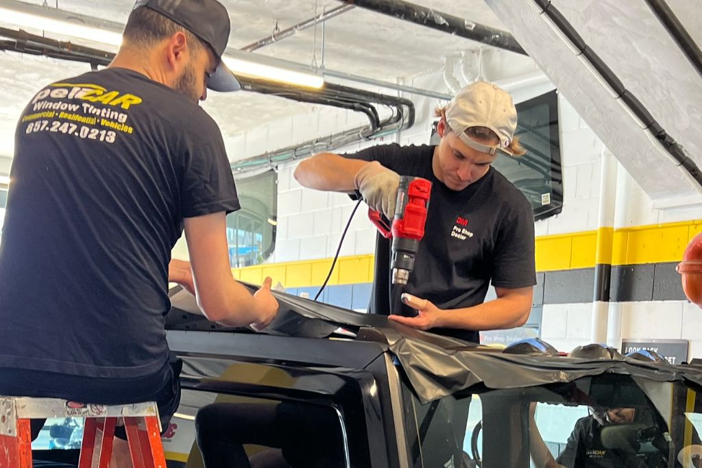 Two Pelicar technicians apply a dark vinyl wrap to a vehicle roof; one uses a heat gun while the other smooths the material. Both wear branded shirts, working efficiently inside a well-lit garage.