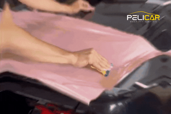 Animated clip of a technician carefully applying a soft pink vinyl wrap to a vehicle’s surface, using a squeegee to smooth out air bubbles. The Pelicar logo appears in the top right corner.