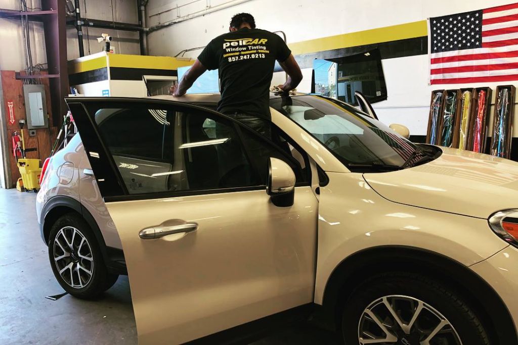 A Pelicar technician works on a cream-colored SUV inside the shop, standing over the roof with the driver’s door open. Rolls of colorful vinyl wrap are visible in the background beside an American flag.