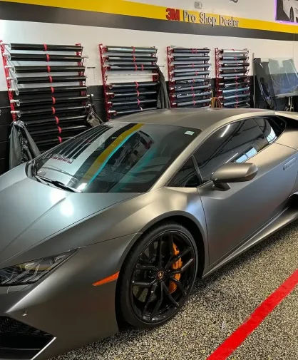A matte gray Lamborghini Huracán parked inside a professional window tinting shop. The car features black wheels, orange brake calipers, and dark tinted windows. Rolls of tint film and a technician working in the background showcase the shop’s setup.