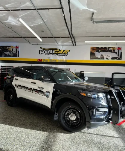 A black and white MIT Police SUV parked inside a professional window tinting shop. The vehicle features a push bumper, steel wheels, and tinted windows. The shop's branding, "peliCAR Window Tinting," is displayed on the wall in the background.