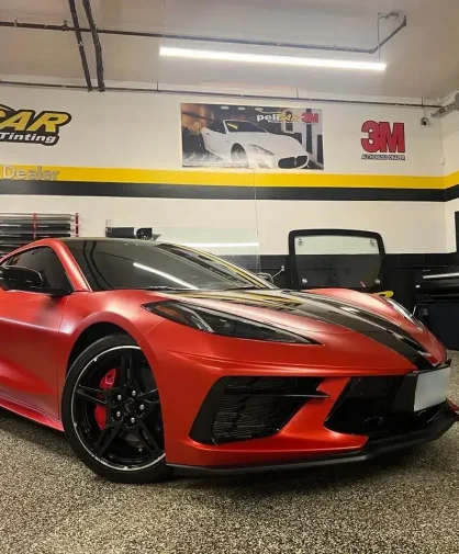 A red Chevrolet Corvette C8 with black racing stripes and tinted windows parked inside a professional window tinting shop. The car features a sleek, aerodynamic design, black wheels with red brake calipers, and an aggressive stance. The "peliCAR Window Tinting" branding and 3M Pro Shop Dealer signage are visible on the walls in the background.
