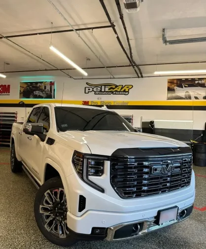 A white GMC Sierra Denali pickup truck parked inside a professional window tinting shop. The truck features a bold black grille, chrome accents, tinted windows, and large multi-spoke alloy wheels. The "peliCAR Window Tinting" branding and 3M Pro Shop Dealer signage are visible on the walls in the background.