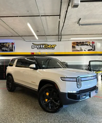 A white Rivian R1S electric SUV parked inside a professional window tinting shop. The vehicle features blacked-out wheels with yellow brake calipers, a sleek modern design, and tinted windows. The shop's branding, "peliCAR Window Tinting," is visible on the wall in the background.