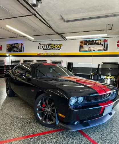 A black Dodge Challenger with red racing stripes parked inside a professional window tinting shop. The muscle car features tinted windows, chrome wheels, red brake calipers, and an aggressive front grille. The "peliCAR Window Tinting" branding and 3M Pro Shop Dealer signage are visible on the walls in the background.