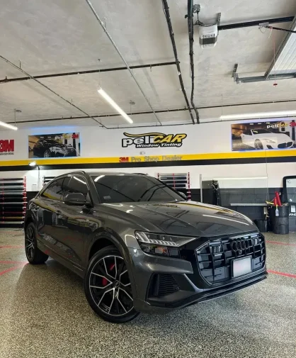 A dark gray Audi SUV with tinted windows parked inside a professional window tinting shop. The vehicle features black alloy wheels with red brake calipers, a large blacked-out grille, and a sleek, sporty design. The "peliCAR Window Tinting" branding and 3M Pro Shop Dealer signage are visible on the walls in the background.